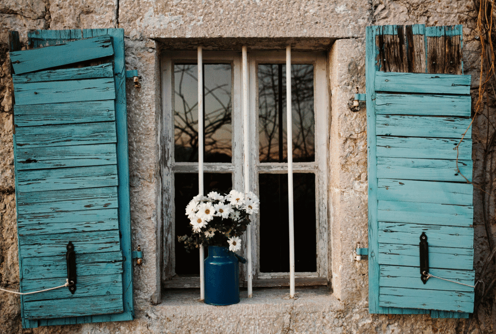 Ventana antigua con persianas de madera en color turquesa y flores blancas en jarrón | Fachada rústica con ventana de madera y persianas azules, decorada con flores blancas en jarrón, ideal para proyectos de instalaciones y reformas tradicionales | Ventana decorativa de estilo rústico con persianas de madera y flores blancas en un jarrón azul, resaltando la belleza de instalaciones tradicionales y reformas con toque vintage en exteriores o interiores.