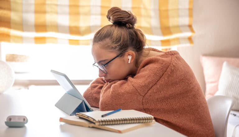 Baja batería en línea, mujer joven con auriculares, usando tableta, en un ambiente de estudio o trabajo en casa, con notebooks y útiles de oficina.